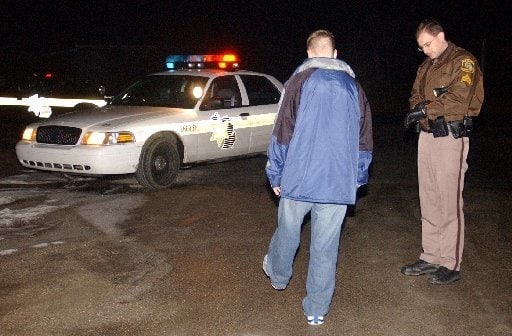 Police officer standing beside a patrol car with flashing lights, interacting with a man taking a one leg stand test.