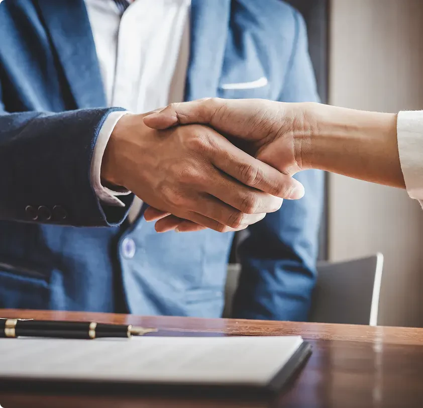 Two people shaking hands over a table with a pen and document, one wearing a blue suit jacket.